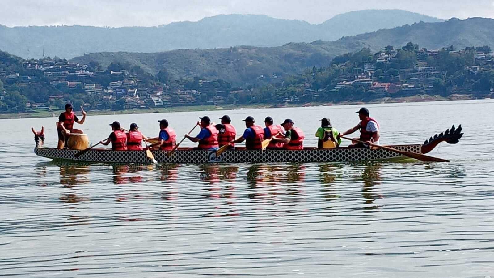 Ein Drachenboot mit Ruderern in roten Schwimmwesten, einem Drachenkopf am Heck und einem ruhigen See mit Hügeln im Hintergrund.