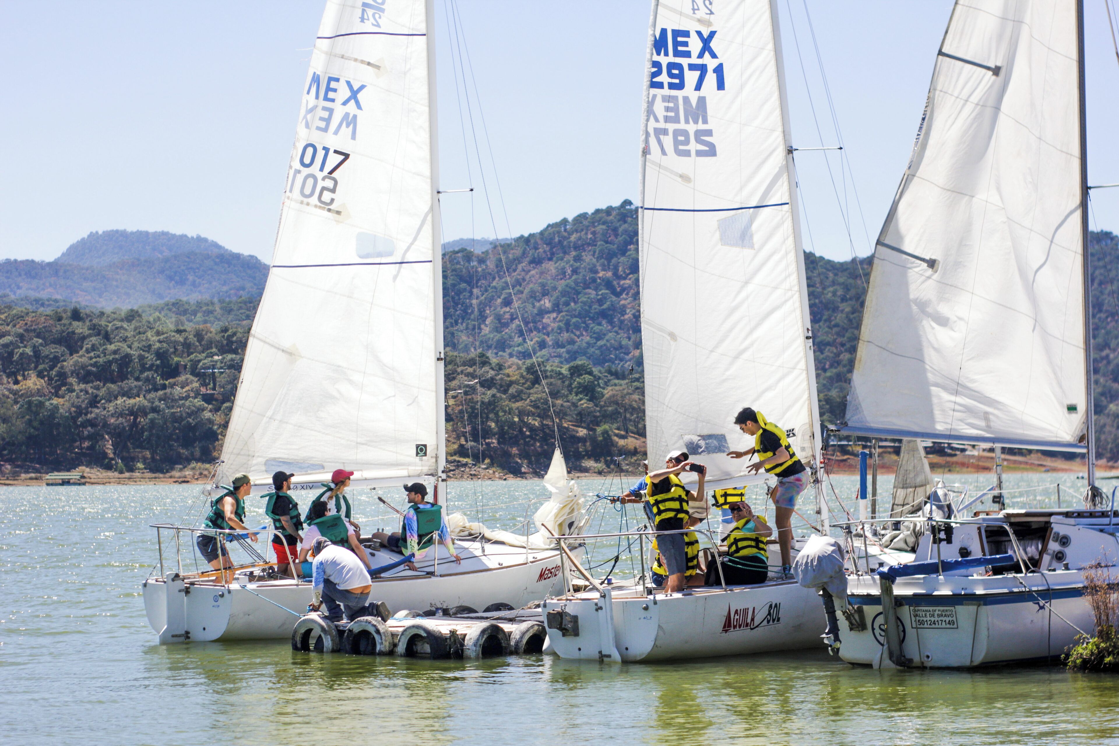 Un grupo de veleros amarrados en una marina, con gente a bordo y un paisaje montañoso al fondo.