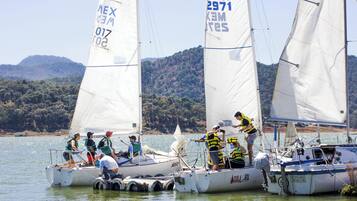 Un grupo de veleros amarrados en una marina, con gente a bordo y un paisaje montañoso al fondo.