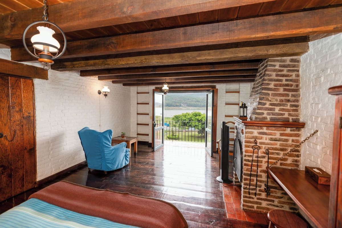 Una habitación con techo de madera, chimenea de ladrillo, una cama, una silla azul y vista a un lago.