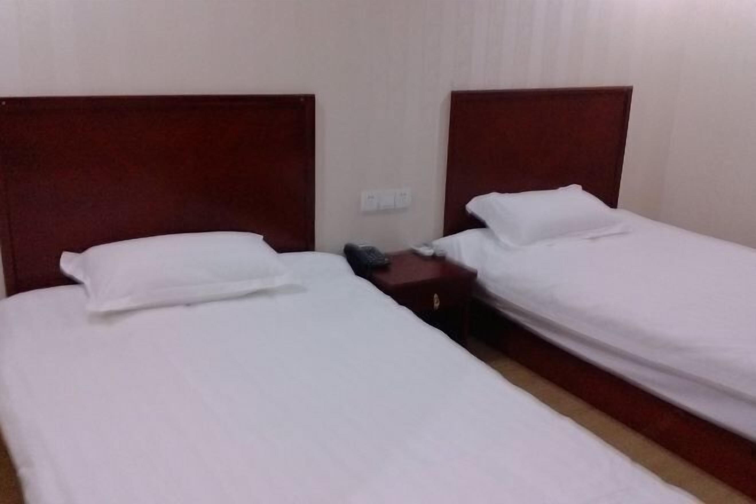 A hotel room with two single beds, wooden headboards, white linens, a small bedside table with a lamp, and a power outlet on the wall.