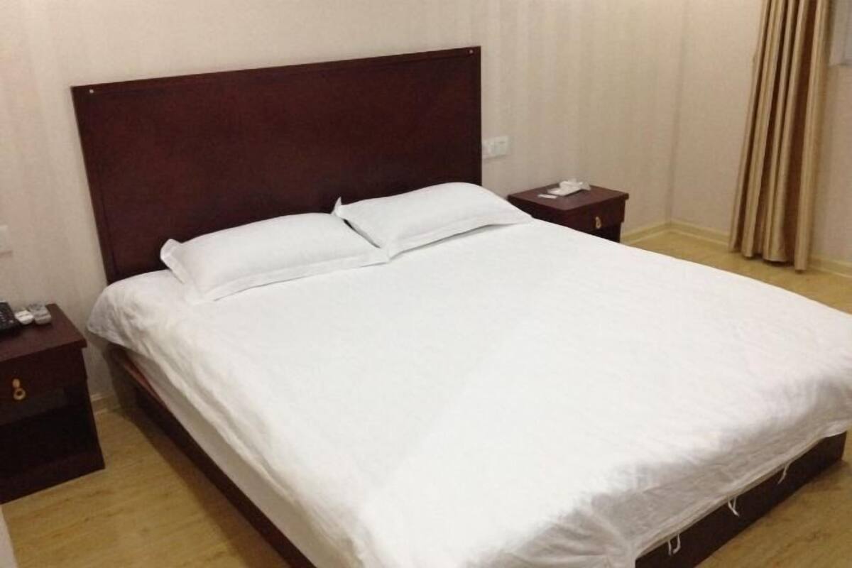 A standard hotel room with a wooden headboard, a single bed with white bedding, a nightstand with a phone and remote control, and a curtain on the window.