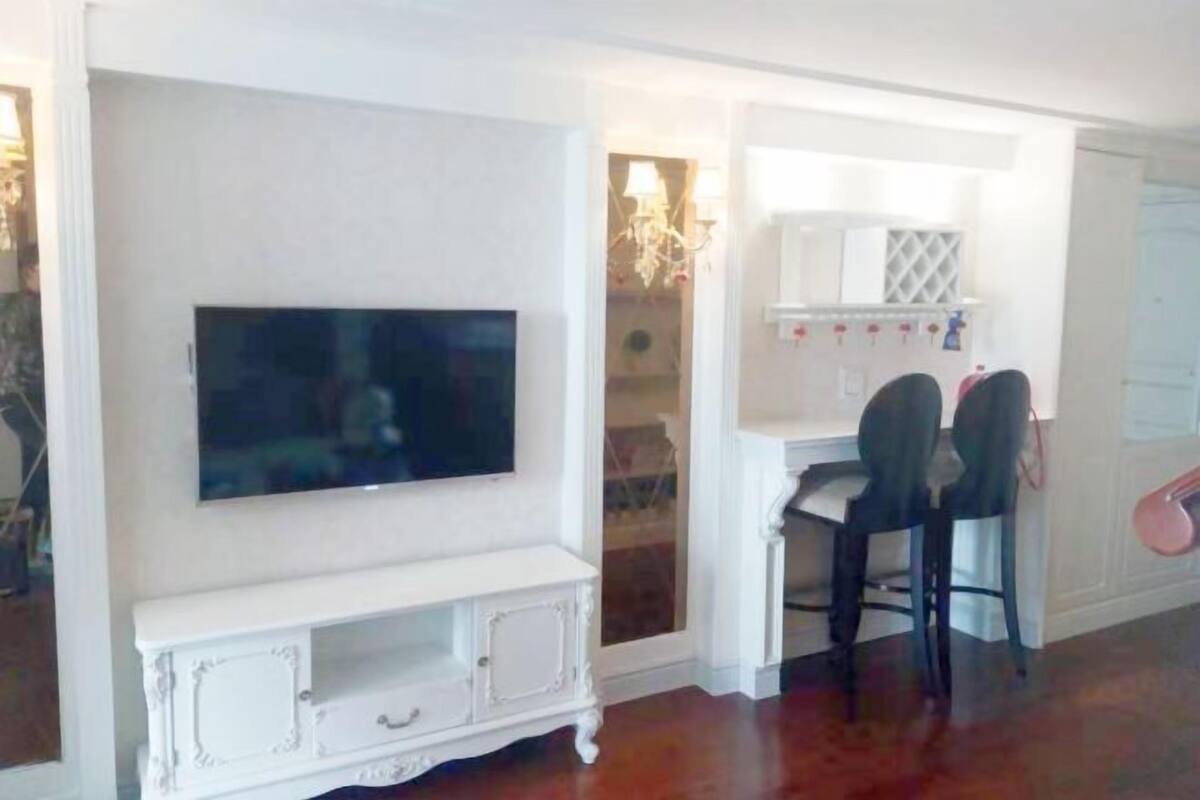 A room with a flat-screen TV mounted on a white cabinet, a small white table, and two black chairs.