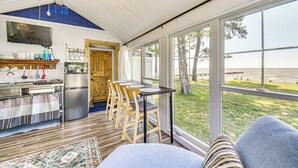 A compact kitchen with modern appliances, a dining area with wooden chairs, and large windows offering a view of the beach. - Tiny House w/ Big Views: Cabin in Shiloh! (Shiloh)