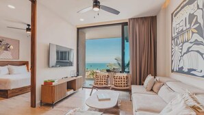 A spacious living area with a sofa, armchairs, a coffee table, and a bed. A large window offers a view of the ocean. - Marina at Caivama (Sayulita)