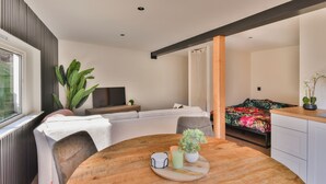 A modern living room with a sofa, a dining table with a plant, and a bed with a colorful bedspread.