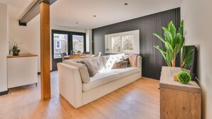 A modern living room with a sofa, wooden flooring, a small table with a plant, and a window with blinds.