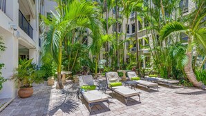 A courtyard with lounge chairs and tables, surrounded by tall palm trees and potted plants.