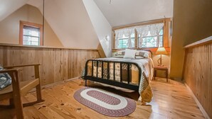 Un dormitorio con techo inclinado, paredes de madera, cama con cabecera, mesita de noche, lámpara y ventana con cortinas.