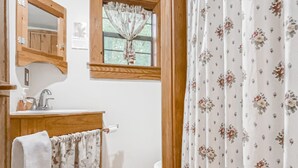 Un baño con piso de madera, un lavamanos con espejo, una ventana con cortina y un inodoro.