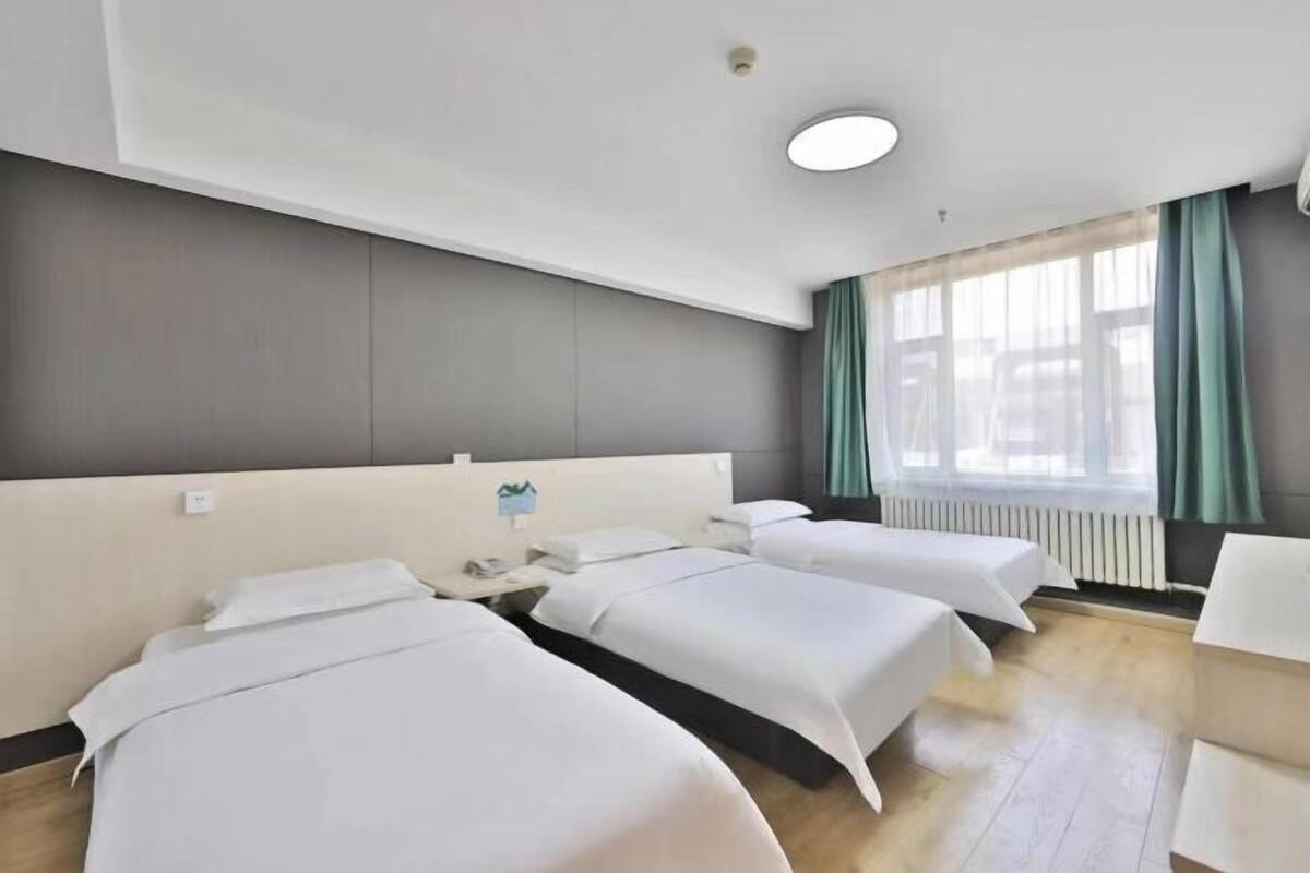 A hotel room with three single beds, a small table, a window with curtains, and a radiator.