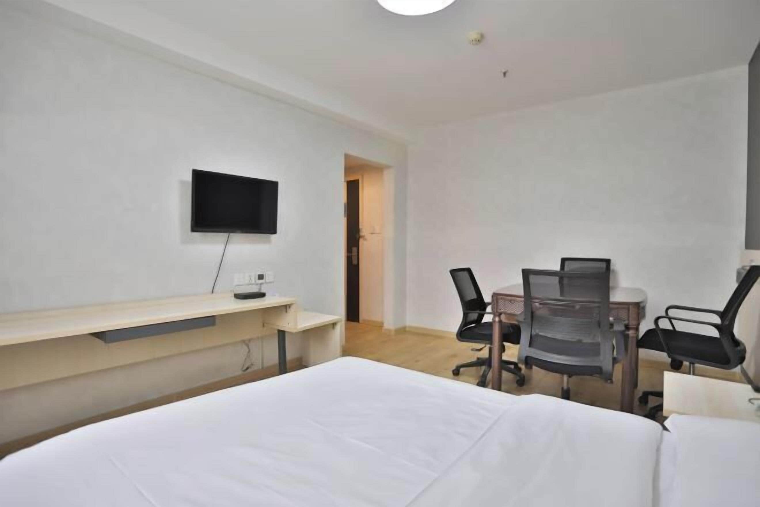 A hotel room with a bed, a desk with a chair, a TV mounted on the wall, and a door leading to another room.