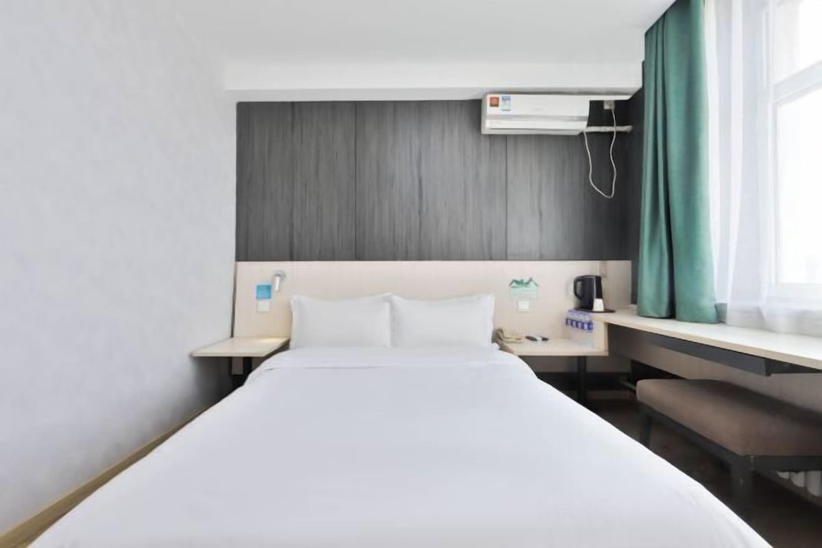 A hotel room with a bed, a desk, a chair, a nightstand, an air conditioning unit, and a window with curtains.