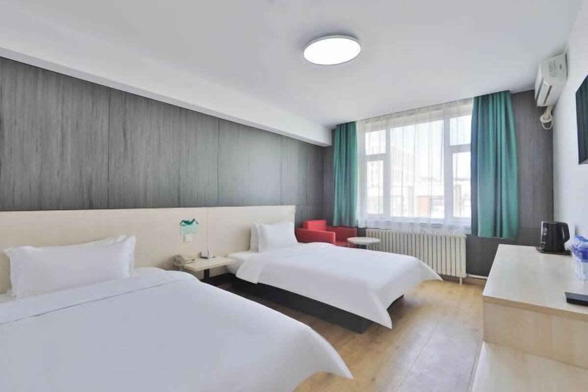 A hotel room with two beds, a desk, a chair, a radiator, and a window with curtains.