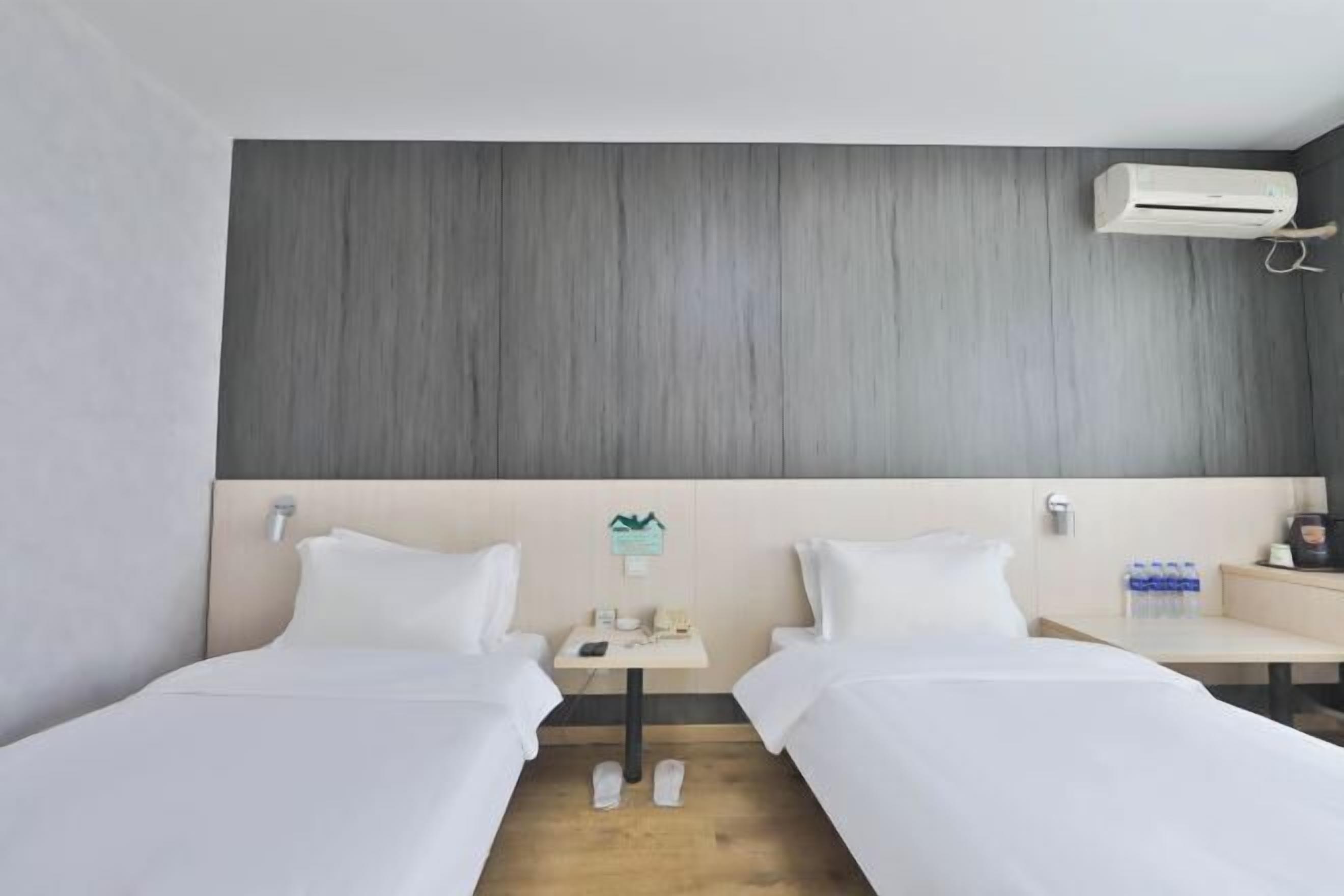 A hotel room with two single beds, a small table with a phone and a lamp, a wall-mounted air conditioner, and a water dispenser on the nightstand.
