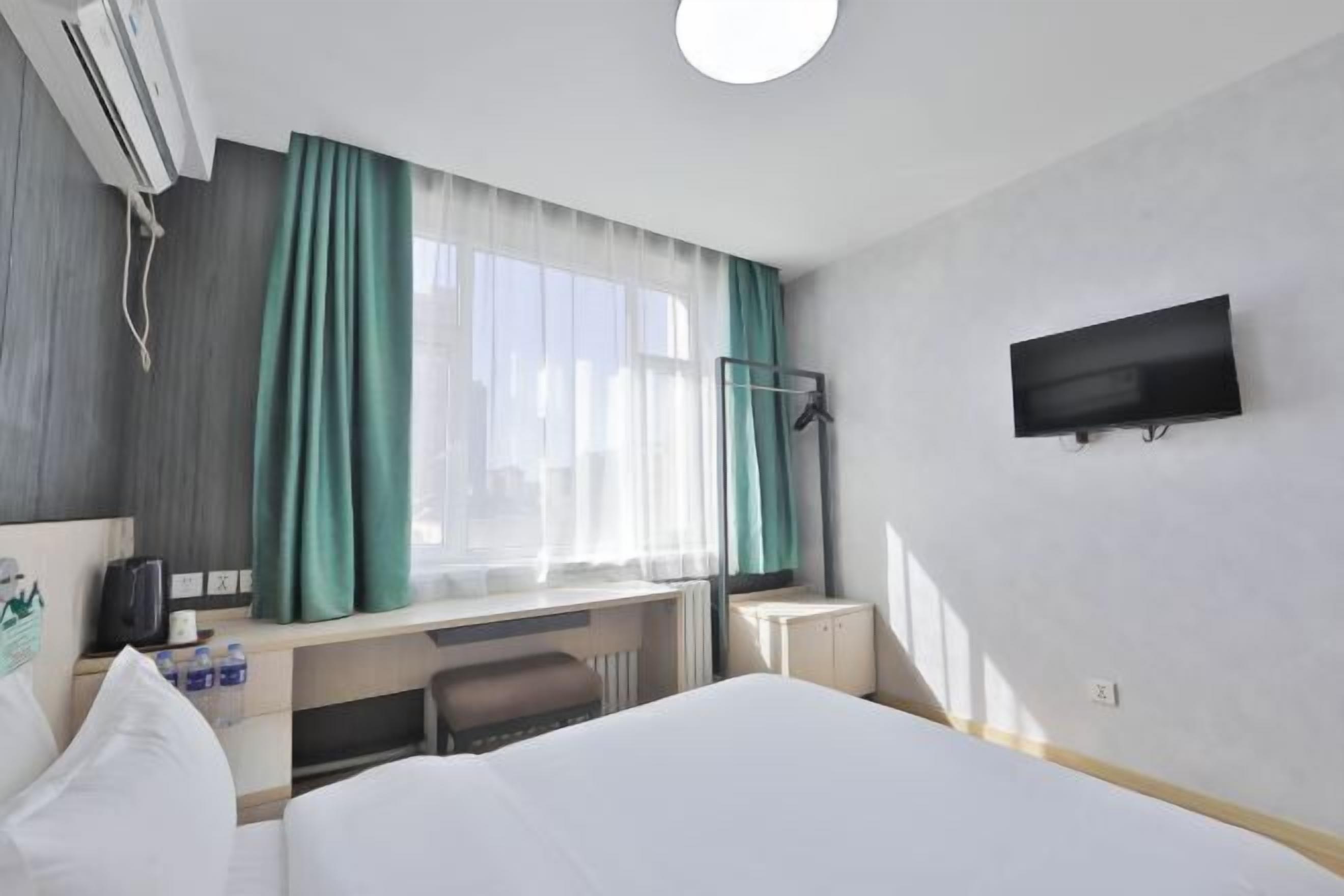 A hotel room with a bed, a desk, a chair, a TV, a window with curtains, and an air conditioning unit.