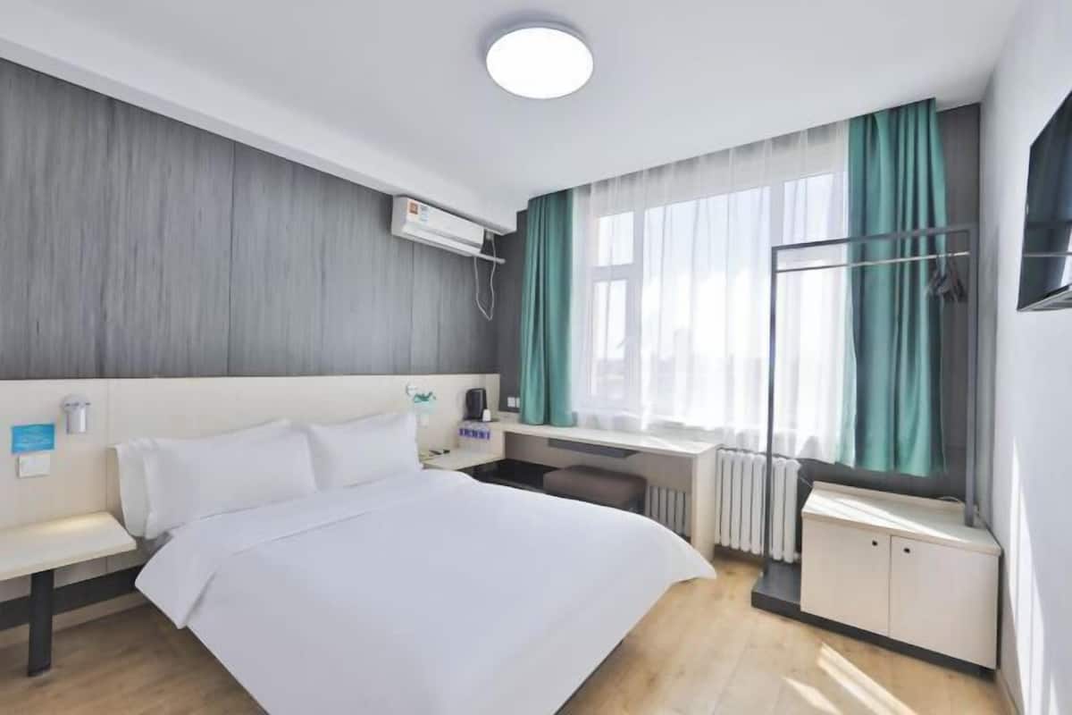 A hotel room with a bed, a desk, a chair, a radiator, and a window with curtains.
