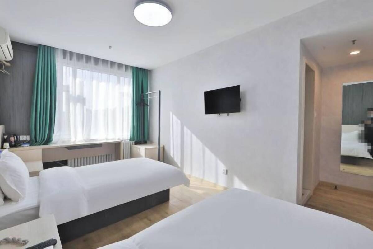 A hotel room with two beds, a TV mounted on the wall, a window with curtains, and a mirror on the wall.