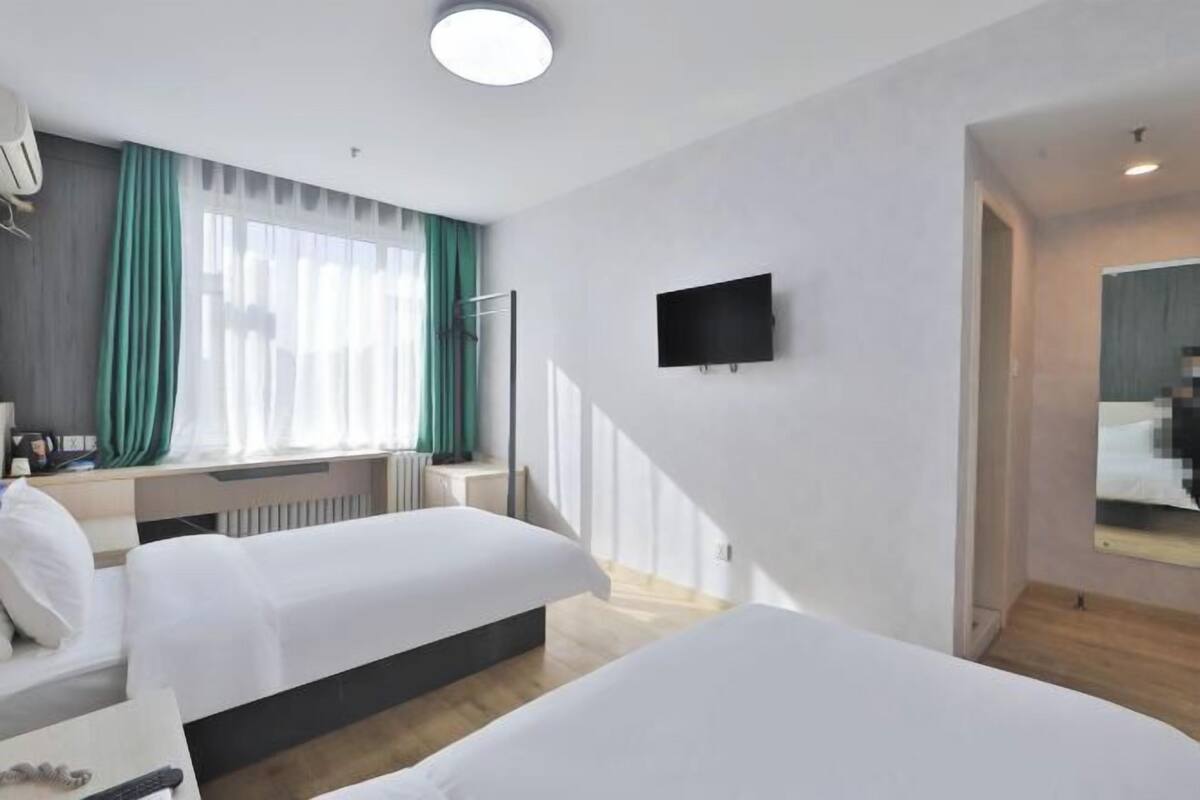A hotel room with two beds, a TV, a desk with a chair, and a large window with curtains.