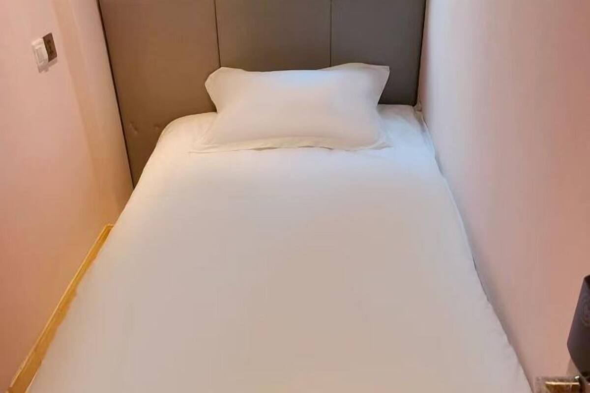 Une chambre d’hôtel étroite, avec un lit simple, une tête de lit et une petite table de chevet.