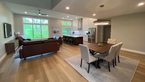 A spacious living room with a large brown sofa, a dining table with four chairs, and a kitchen area with modern appliances.