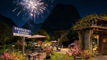 Un restaurante con mesas al aire libre, rodeado de montañas y un cielo nocturno con fuegos artificiales.