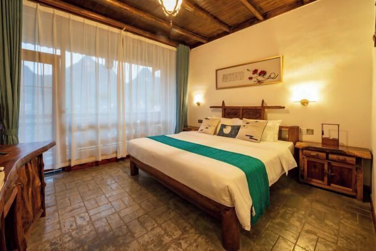 Quarto com cama, cabeceira de madeira, mesas de cabeceira, um criado-mudo, janela com cortinas e um quadro pendurado na parede.