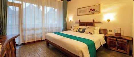 Un dormitorio con una cama, cabecera de madera, mesitas de noche, una mesita auxiliar, una ventana con cortinas y un cuadro colgado en la pared.