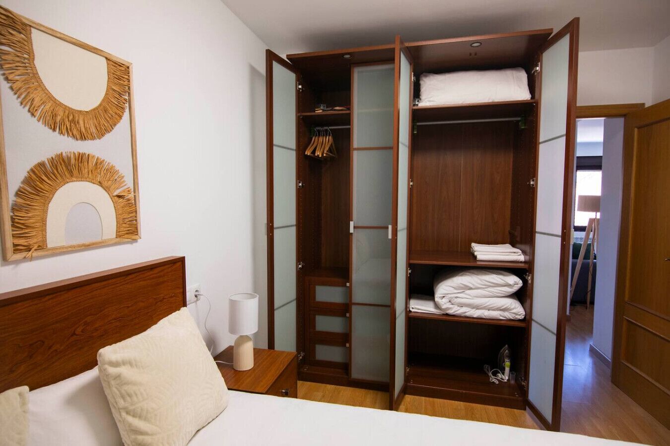 A bedroom with a bed, a wooden wardrobe with a mirror, and a nightstand with a lamp.