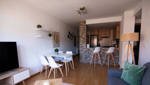 Un salon moderne avec un canapé, une télévision, une table à manger et des chaises. On aperçoit une cuisine au fond.