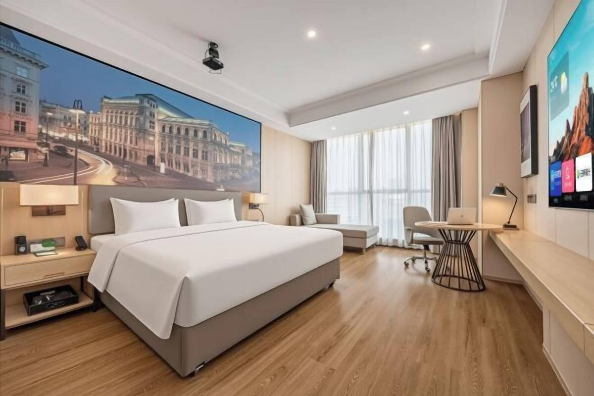 A hotel room with a large bed, a desk, a chair, a TV, and a large window with curtains.
