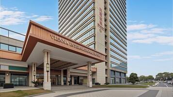 Un hotel moderno con una entrada destacada, un edificio alto al fondo y un cielo despejado.