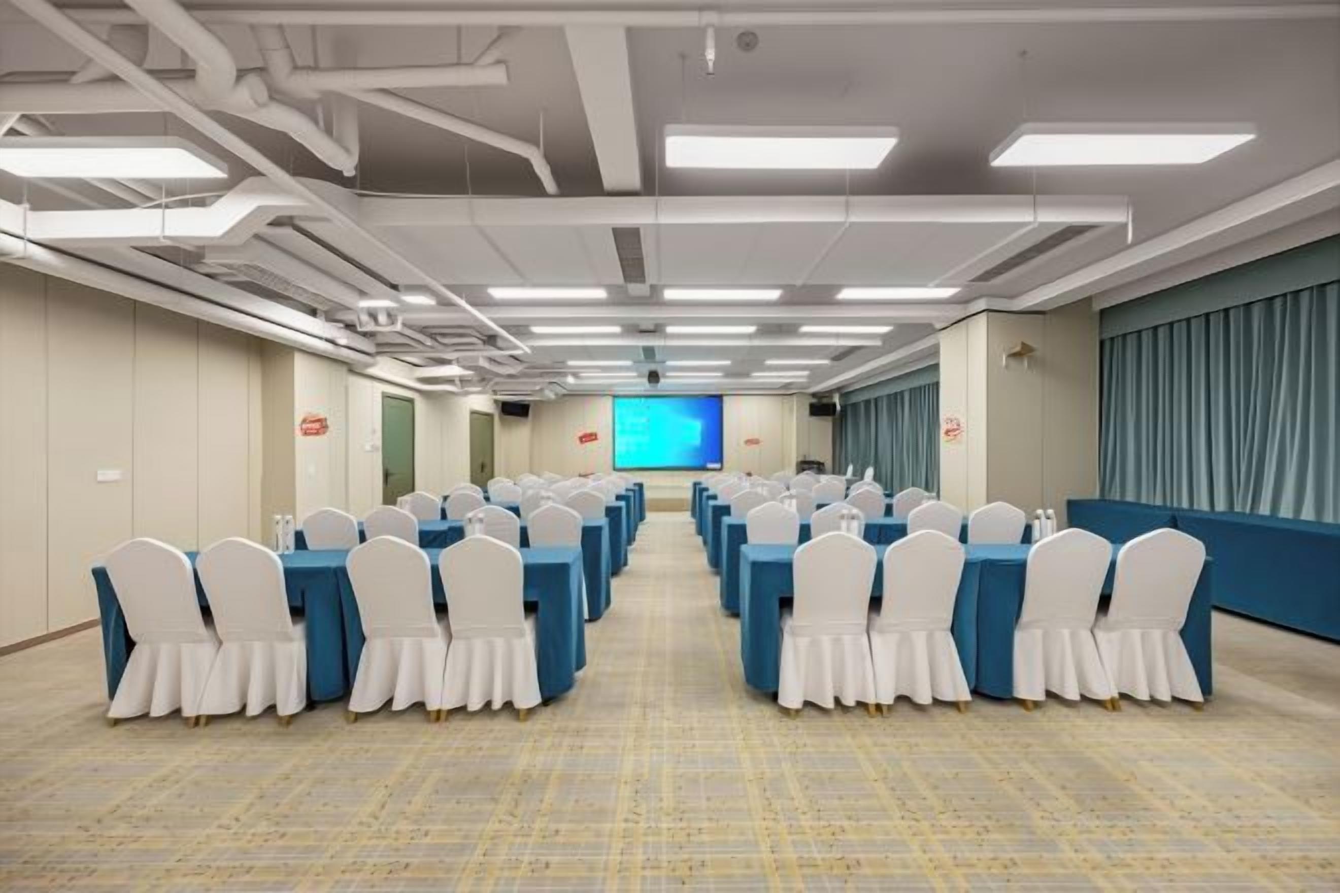 Un salón de conferencias con filas de mesas y sillas, una pantalla grande al frente y una pared azul de acento.