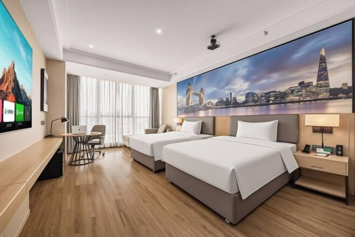 A hotel room with two beds, a desk, a chair, a TV, and a large cityscape painting on the wall.