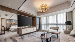 A modern hotel room with a large bed, a sofa, a coffee table, a TV, and a large window with curtains.