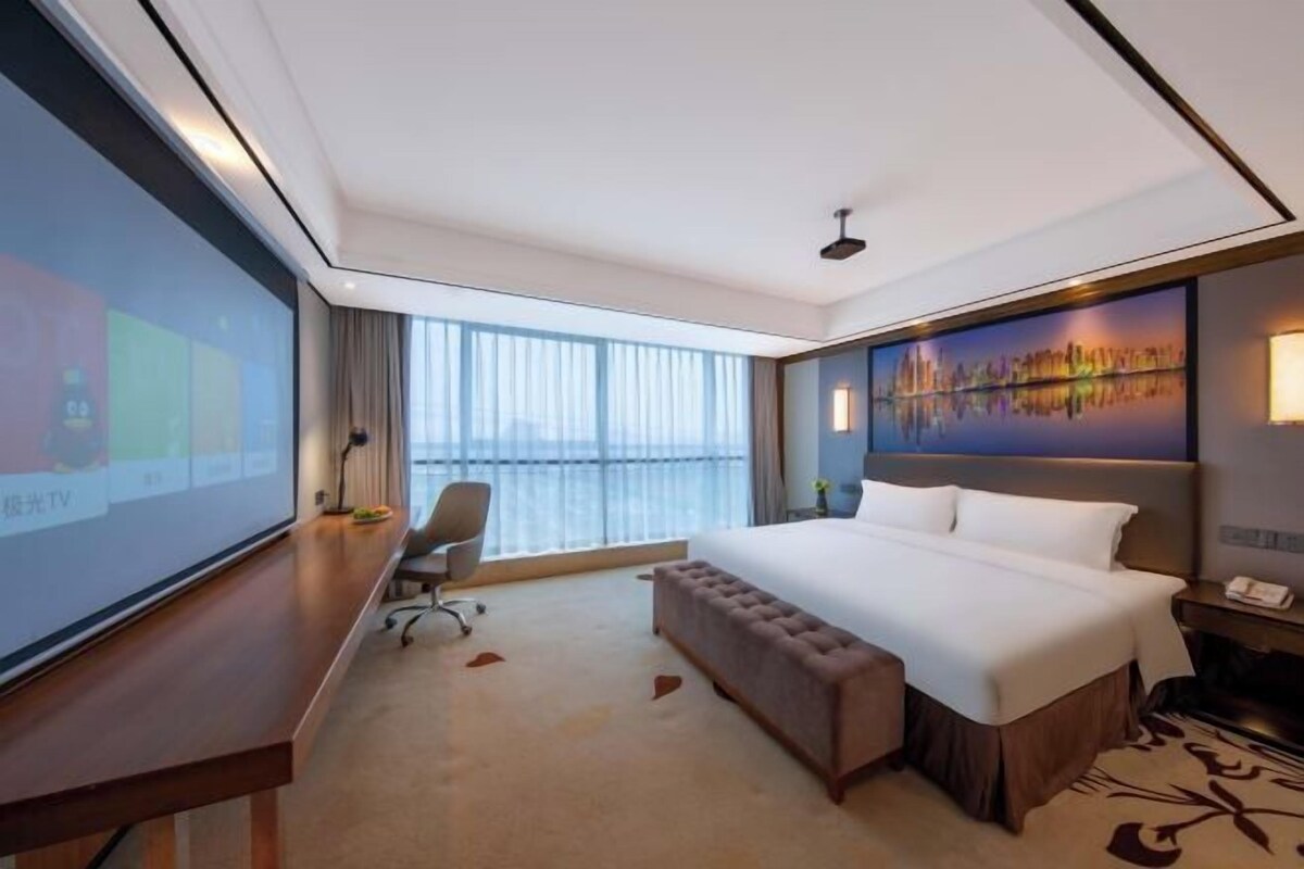 Ein Hotelzimmer mit einem großen Bett, einem Schreibtisch mit Fernseher, Blick auf die Stadt und einem Balkon.