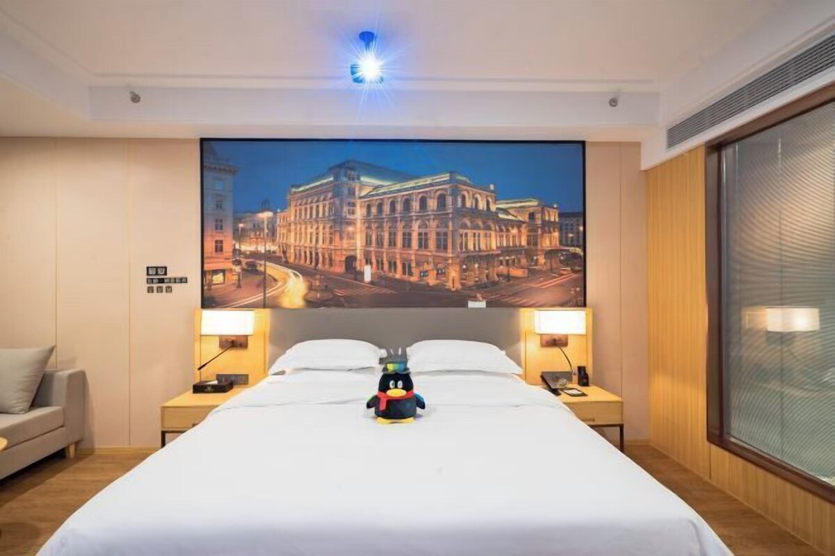 Ein Hotelzimmer mit einem großen Bett, zwei Nachttischen inklusive Lampen, einer weichen Pinguin-Spielzeugfigur auf dem Bett und einer Wandmalerei mit Stadtansicht.