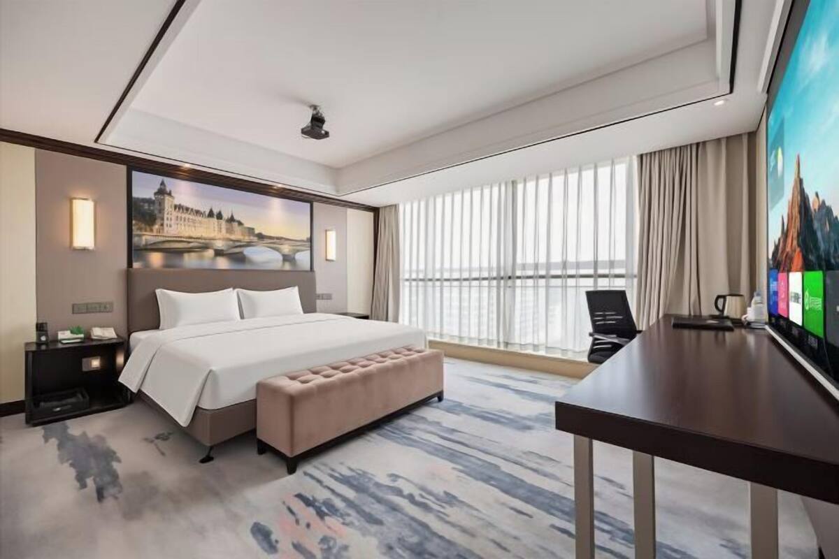 A modern hotel room with a large bed, a desk, and a view of a cityscape.
