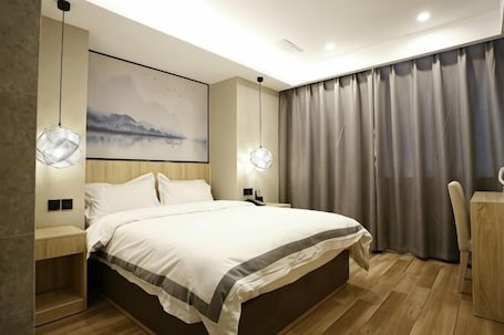 A modern bedroom with a large bed, bedside tables, a desk, and a view of a cityscape through curtains.. Qingmu Hotel (Nanjing Xinzhuang Metro Station)