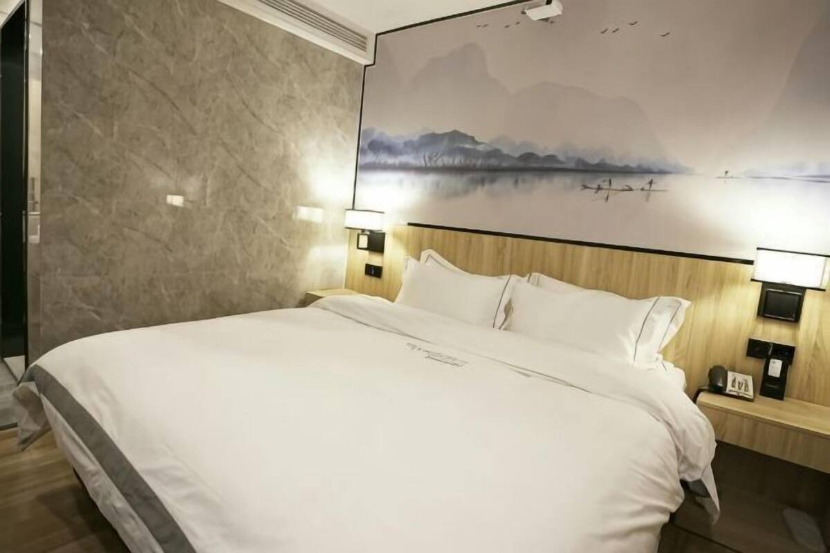 A hotel room with a large bed, wooden headboard, two bedside tables with lamps, a wall-mounted TV, and a scenic mural.