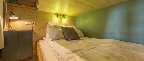 A bedroom with a wooden ceiling, a bed with white bedding, a nightstand with a lamp, and a green wall.