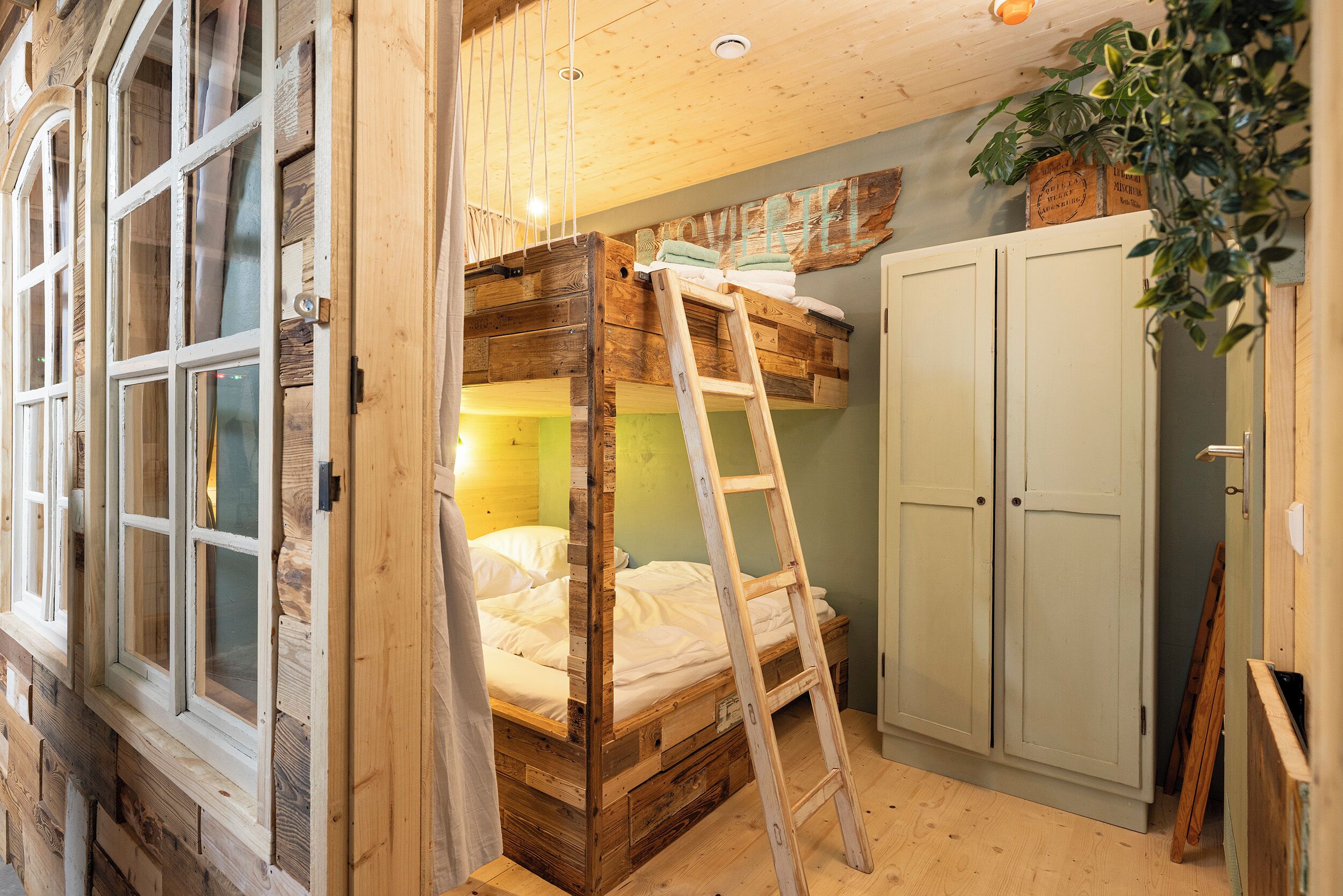 A rustic room with a loft bed, a ladder, a wardrobe, and a window with curtains.
