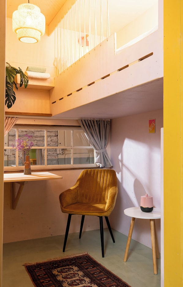 A room with a yellow chair, a small table with a pink container, a window with curtains, a hanging light fixture, and a plant on a shelf.
