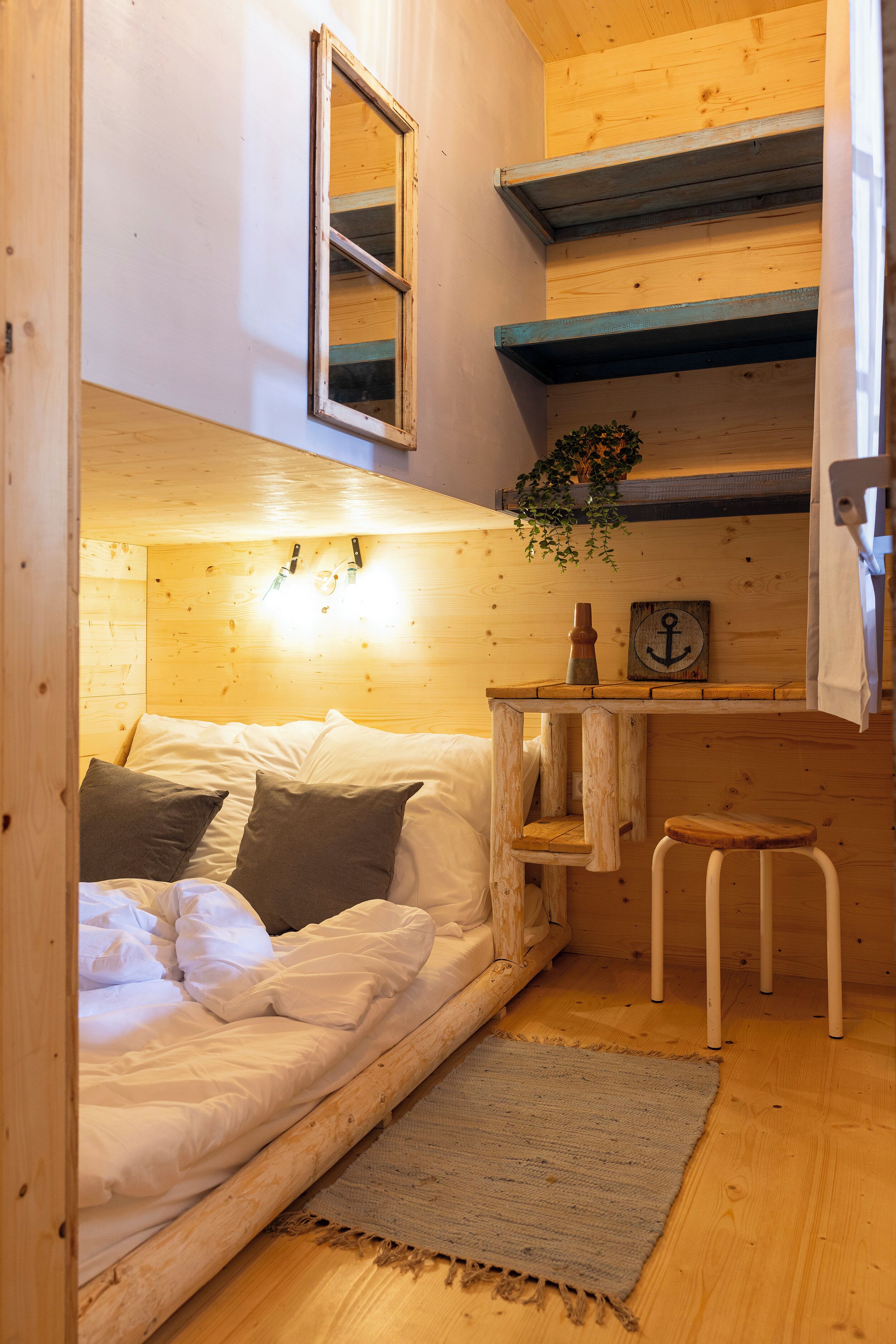 A cozy room with a bed, a small table with a plant, a stool, and a wooden shelf with a mirror and an anchor decoration.