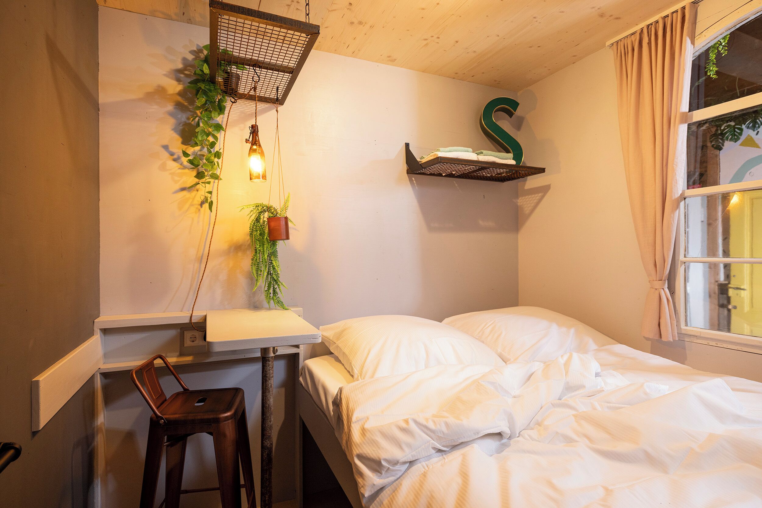 A bedroom with a bed, a desk, a chair, a hanging plant, a shelf with a letter, and a window with curtains.
