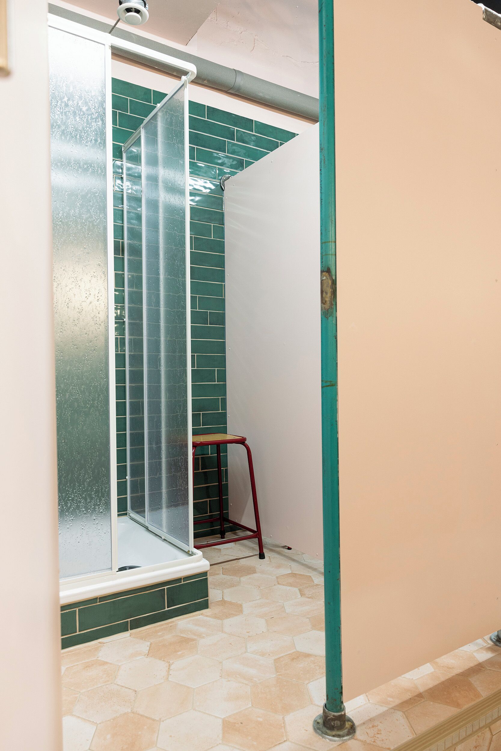 A shower with a glass door, a red stool, and a green tiled wall.