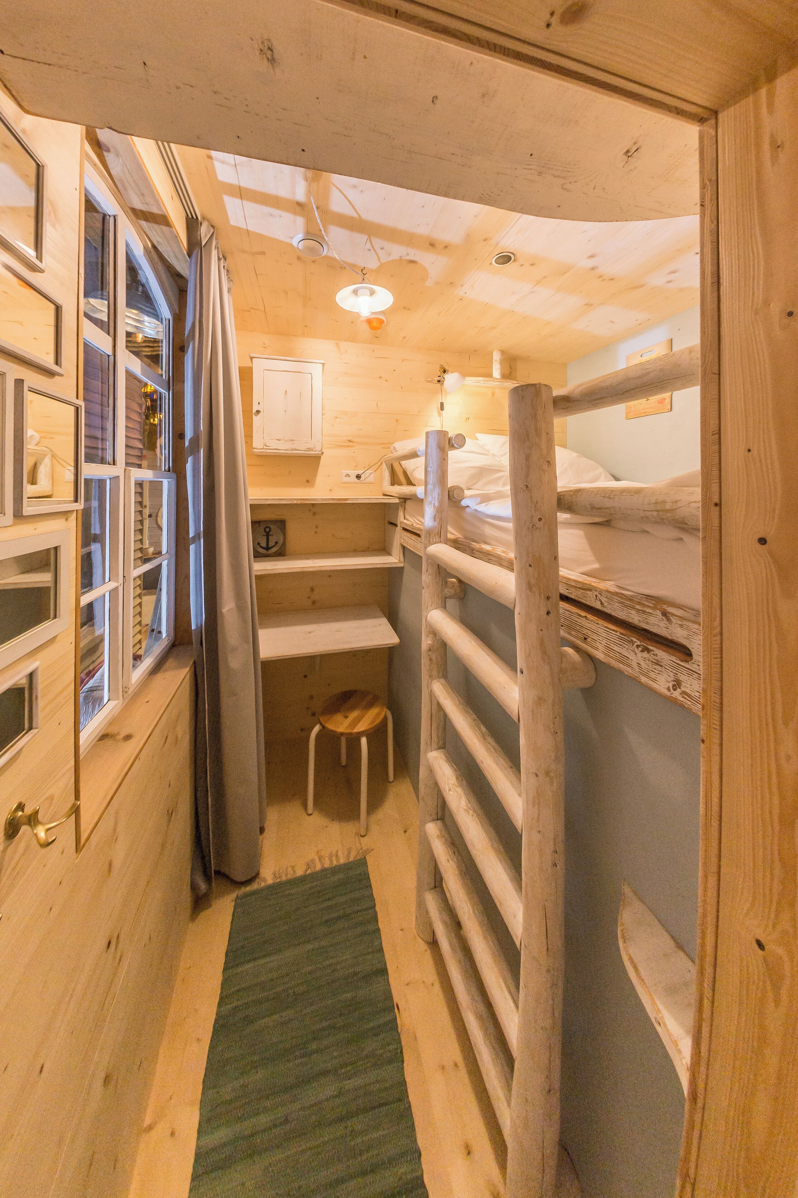 Une petite pièce en bois avec des lits superposés, un petit tabouret, une fenêtre avec des rideaux et un tapis vert.