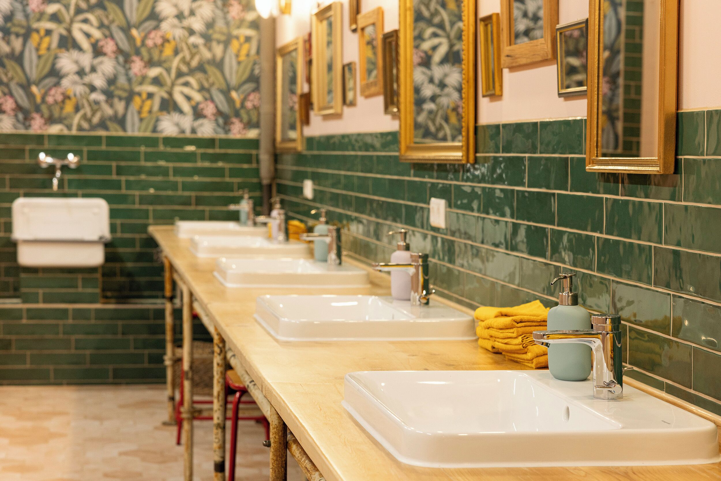 Uma fileira de pias brancas com torneiras cromadas, paredes revestidas de azulejos verdes e espelhos emoldurados acima de cada pia.