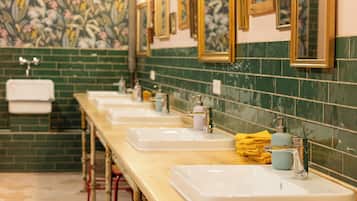 Uma fileira de pias brancas com torneiras cromadas, paredes revestidas de azulejos verdes e espelhos emoldurados acima de cada pia.