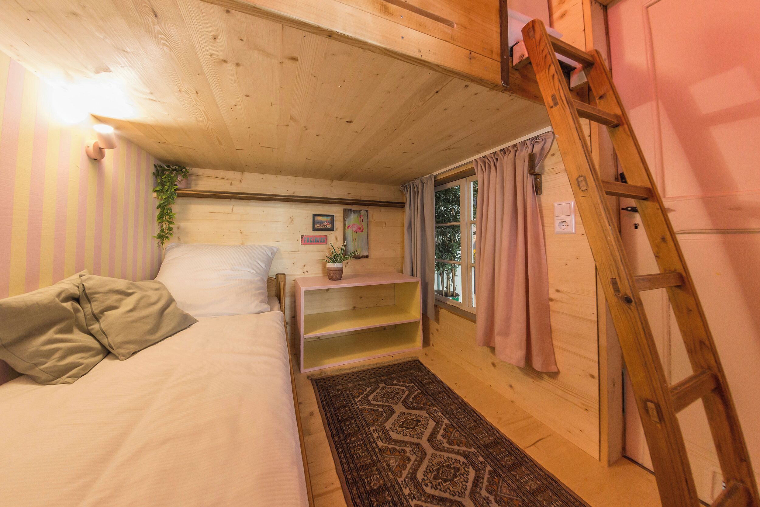 Une chambre à coucher accueillante, avec un lit, une échelle en bois, une étagère avec une plante et une fenêtre avec des rideaux.